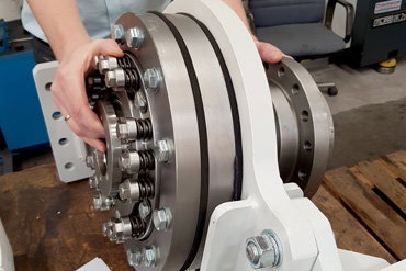 Eine Person hält ein großes metallisches Bauteil mit mehreren Schrauben und Ringen, das auf einer weißen Halterung montiert ist. Im Hintergrund sind Werkstattmöbel und Geräte zu sehen.